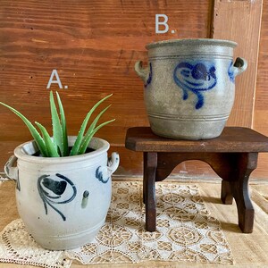 Op de afbeelding: Twee antieke stenen potten, een met een groene plant, de andere leeg, staan op een kleine houten tafel. De potten hebben blauwe bloemmotieven en handvatten. De achtergrond is een houten deur.