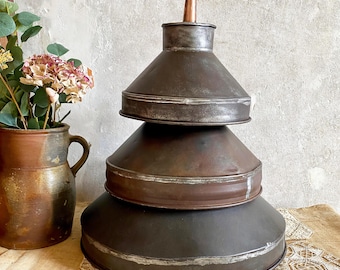May include: A stack of three graduated dark metal funnels with a weathered patina, topped with a small copper-colored spout. The funnels are arranged in a tiered, conical shape, showcasing a rustic, industrial aesthetic with visible metallic textures.