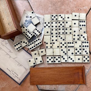 Peut inclure: Un jeu de dominos blanc cassé avec des points noirs, disposés en grille et en tas épars. Comprend une boîte de rangement rectangulaire en bois, un couvercle en bois et une enveloppe de style ancien avec du texte cursif et un timbre rouge.