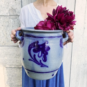 Puede incluir: Una maceta grande de cerámica azul grisáceo con dos asas y un diseño floral azul oscuro. La maceta está llena de vibrantes flores magenta intenso. La estética general es rústica y encantadora.