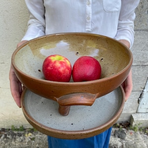 May include: A handmade ceramic fruit bowl with a two-tone design, holding two red apples. The bowl has a brown exterior and a speckled beige interior. The bowl is supported by a matching base.