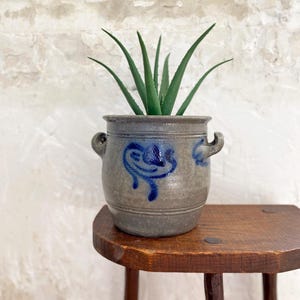 Könnte beinhalten: Ein grauer Steinguttopf mit zwei seitlichen Griffen und einem blauen, kobaltfarbenen, schlickerbemalten Blumenmuster auf der Vorderseite. Das zylindrische Gefäß enthält eine grüne Aloe-Vera-Pflanze mit langen, spitzen, gezackten Blättern.