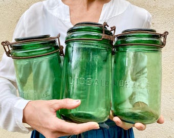 May include: Three vintage green glass jars with wire bail closures. Each jar features the embossed text L'IDEALE on the front. The glass is translucent and tinted green, paired with rusted metal hardware for securing the lids. These storage containers.