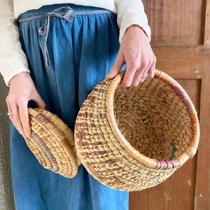 Può includere: Due cesti intrecciati dall'aspetto naturale e strutturato. Il cesto più grande è rotondo con un'ampia apertura e un bordo colorato. Il cesto più piccolo è un coperchio abbinato. I cesti sono tenuti contro una gonna blu e un top bianco.