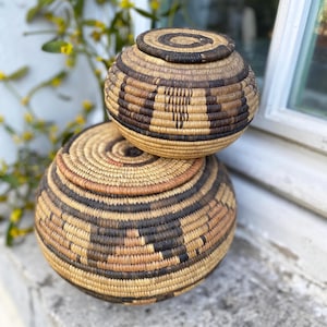 May include: Two round, lidded baskets made of woven natural fibers. The baskets feature alternating bands of light and dark brown, creating geometric patterns. The lids fit snugly on top. The baskets are likely used for storage or decoration.