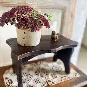 May include: A small, dark brown wooden stool with a decorative top. A ceramic pot with burgundy flowers and a small bronze bird figurine sit on the stool. The stool is placed on a wooden surface with a lace doily.