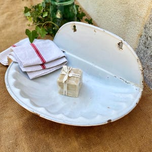 May include: A white enameled metal wall-mounted basin with a scalloped edge, featuring a stack of white linen towels with red stripes and a square bar of soap wrapped in paper with the text "LE SAVON DE PROVENCE" printed on the label.