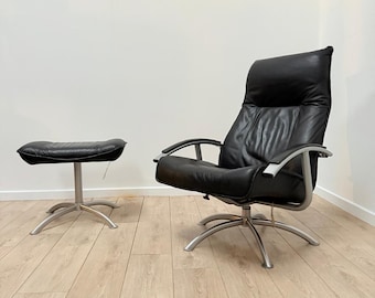 May include: Black leather armchair and matching ottoman set. The chair has a high back, curved armrests, and a silver metal frame. The ottoman is a small, round, black leather cushion on a silver metal base. The set is on a light wood floor.
