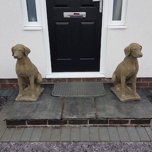 May include: Two brown stone dog statues sit on a stone step in front of a black door. The statues are facing forward and have their heads tilted slightly to the side. The door is surrounded by white trim and the step is made of stone.