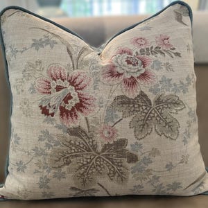 May include: Square decorative pillow with a floral pattern in shades of pink, burgundy, and gray on a cream background. The pillow has a teal blue trim around the edges. The floral design includes large flowers and detailed leaves.