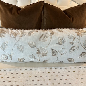 May include: Two brown velvet pillows with a white pillow with a floral pattern in the center. The floral pattern is in shades of brown and silver.