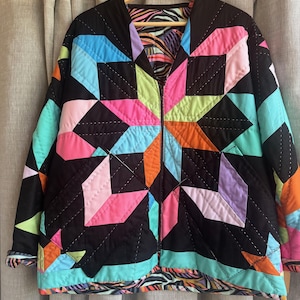 May include: A quilted jacket with a vibrant geometric pattern in shades of pink, blue, green, orange, and purple on a black background. The jacket has a V-neck and a zipper closure. The lining features a colorful abstract design.