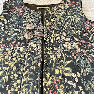 May include: A sleeveless vest with a floral print featuring a variety of flowers and leaves in shades of green, yellow, red, and white. The vest has a brown button closure with a brown cord.