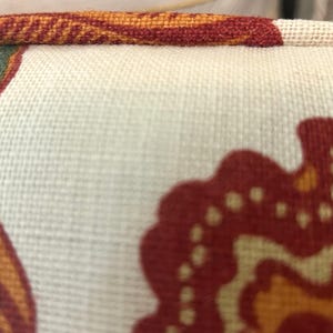 Może przedstawiać: Zbliżenie na poduszkę z teksturowanej tkaniny w kolorze złamanej bieli, ozdobioną wzorem z organicznymi kształtami w odcieniach palonej pomarańczy, musztardowej ż&oacute;łci i szałwiowej zieleni. Krawędź poduszki wykończona jest grubą, pasującą lam&oacute;wką.