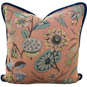May include: A square throw pillow featuring a salmon-pink background with a botanical print of lotus flowers, seed pods, and various leaves in shades of green, yellow, and blue. The pillow is finished with a dark navy blue piped edge trim.