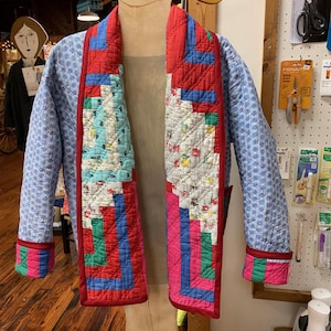 May include: A quilted jacket displayed on a mannequin. The jacket features a blue patterned body with red trim and colorful patchwork detailing on the lapels and cuffs. The jacket is open, revealing the interior.