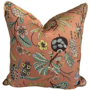 May include: A square decorative pillow with a peach background and a botanical print featuring flowers, leaves, and seed pods in shades of pink, yellow, and blue. The pillow has a fringed edge.
