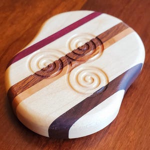 Könnte beinhalten: Ein Untersetzer aus Holz mit einem geschnitzten keltischen Knotendesign. Der Untersetzer besteht aus verschiedenen Holzarten, darunter Ahorn, Walnuss und Kirsche. Der Untersetzer hat die Form einer Träne und eine glatte, polierte Oberfläche.