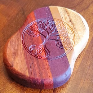 May include: A wooden coaster featuring a tree of life design carved into the surface. The coaster is crafted from various types of wood, including red, brown, and light brown.