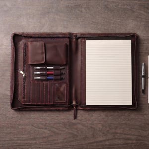 May include: A dark brown leather zippered portfolio open to reveal a lined notepad on the right side. The left interior features a zippered pocket, a flap pocket, three pen loops holding silver, red, and blue pens, and several card slots.
