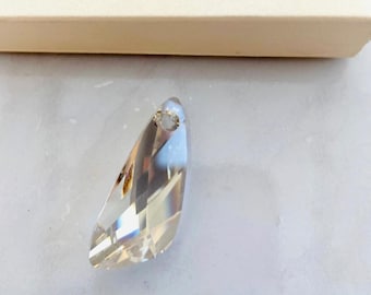 May include: A clear crystal pendant with a teardrop shape and a hole at the top for stringing. The pendant is on a white surface. The text "SWAROVSKI" is visible on the box.