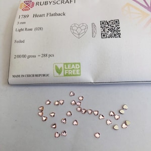 Puede incluir: Un conjunto de pedrería plana en forma de corazón de color rosa claro, de 3 mm, dispersa sobre una superficie blanca. El embalaje dice "RUBYSCRAFT 1789 Heart Flatback Light Rose (028) Foiled". El paquete también incluye un código QR y el texto "LEAD FREE".