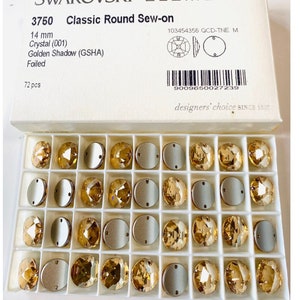 May include: A box of 72 gold-toned Swarovski Elements sew-on crystals, each measuring 14 mm. The crystals are arranged in a grid pattern in a white plastic tray. The box is labeled "Swarovski Elements 3750 Classic Round Sew-on 14 mm Crystal (001) Golden Shadow (GSHA) Foiled 72 pcs 103454356 QCD-TNE M 9009650027239 designers' choice SINCE 1895"