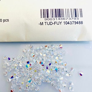 Puede incluir: Primer plano de un montón de cuentas de cristal transparentes. Las cuentas son facetadas y tienen un brillo iridiscente. Las cuentas están empaquetadas en una bolsa blanca con un código de barras y el texto "9003148673793 -M TUD-FUY 104379488".