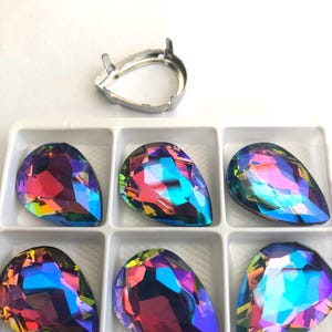 Puede incluir: Seis cristales iridiscentes en forma de lágrima en una bandeja blanca, con un engaste de metal plateado encima. Los cristales muestran una vibrante gama de colores, incluyendo azules, morados y verdes, reflejando la luz.