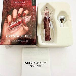 Puede incluir: Una pequeña botella transparente llena de cristales de purpurina rojos. La botella está etiquetada con "Crystal Pixie" y "Heart's Desire". La botella está empaquetada en una caja blanca con el logotipo de Swarovski y el texto "Create Your Style".