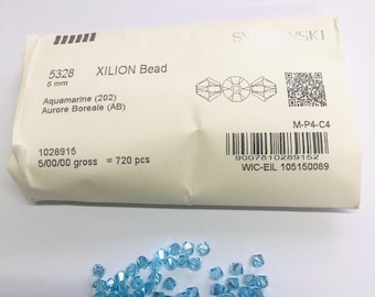 May include: A collection of small, faceted, light blue aquamarine Xilion beads with an Aurore Boreale finish, accompanied by a white paper packaging envelope labeled with the Swarovski brand name, 5328 model number, 5 mm size, and 720 pieces.