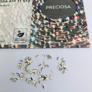 May include: Small, silver-coloured, oval-shaped rhinestones scattered on a white surface. A card with the word "PRECIOSA" and the text "LEAD-FREE BRILLIANCE" is in the background.