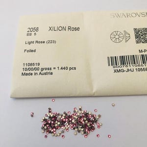 Op de afbeelding: Een stapel lichtroze XILION Rose kristallen, maat SS 5, met een folie achterkant, verspreid op een wit oppervlak. De kristallen liggen naast een verpakking met productinformatie, inclusief de tekst "Light Rose (223)" en "Made in Austria."