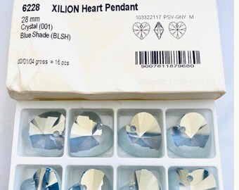 May include: A package of 6 light blue crystal Xilion heart pendants, each measuring 28 mm. The package is labeled with the Swarovski Elements logo and product information, including the product number 6228, the crystal color (001), and the blue shade (BLSH).