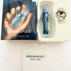 Puede incluir: Un pequeño frasco de vidrio transparente lleno de purpurina azul, etiquetado con "CRYSTALPIXIE™ NAIL ART" y "OCEAN DREAMS PETITE". El frasco está empaquetado en una caja blanca con un fondo azul y negro, que muestra una mano sosteniendo el frasco y el texto "SWAROVSKI CREATE YOUR STYLE" y "CRYSTALPIXIE™ NET WT. 0.18 oz./5g".