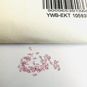 Op de afbeelding: Een strooiing van kleine, regenboogkleurige, roze strasssteentjes in marquise-vorm op een wit oppervlak.
