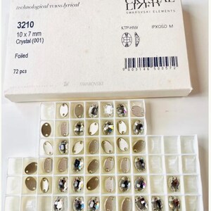 Può includere: Un contenitore di plastica trasparente con 72 piccoli cristalli ovali trasparenti. I cristalli sono disposti in file e colonne. Il contenitore è etichettato con "3210 10 x 7 mm Crystal (001) Foiled 72 pcs" e "CRYSTAL LIZES SWAROVSKI ELEMENTS".
