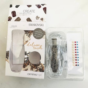 Puede incluir: Un recipiente de plástico transparente lleno de pequeños cristales de color plateado. El recipiente está etiquetado como "Swarovski Deluxe Rush Crystal Pixie". El paquete también incluye una pequeña hoja de tela blanca con una fila de cristales de colores.