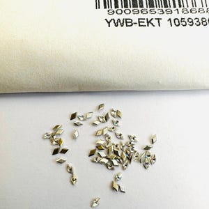 Puede incluir: Una dispersión de pequeños diamantes de imitación plateados en forma de rombo con una superficie reflectante. Algunos diamantes de imitación tienen un centro transparente e iridiscente. Un papel blanco con un código de barras y texto está en el fondo.