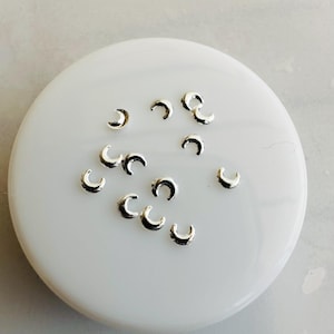 May include: Small, silver crescent moon charms scattered on a white, circular surface. The charms are evenly spaced and reflect light, creating a subtle shine. These charms are ideal for jewellery making or craft projects.