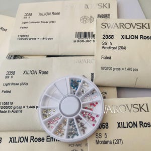 May include: A collection of Swarovski crystals in various colors, including pink, blue, and silver, are displayed in a clear, circular organizer. Several packages with the Swarovski logo and product information are visible in the background.