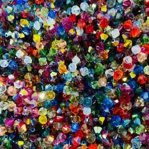 May include: A close-up of a collection of colourful, faceted beads. The beads are a mix of red, blue, green, yellow, and purple, with an iridescent finish. The beads are scattered, creating a textured, eye-catching display.