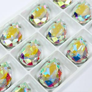 May include: A close-up of a clear plastic container filled with iridescent teardrop-shaped rhinestones. The rhinestones are arranged in rows and have a rainbow-like sheen.