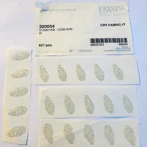 Peut inclure: Un gros plan sur une carte blanche avec le texte "CRYSTAL LIZR SWAROVSKI ELEMENTS CRY.FABRIC-IT 320054 010001AB- CGB-Artik el 627 pcs 800587323 985465 ET14 LAY MADE IN AUSTRIA www.SWAROVSKI.COM CRYSTALLIZED" imprimé dessus. La carte est entourée de petits strass blancs ovales.