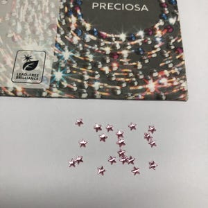 May include: A collection of small, light pink star-shaped crystals scattered on a surface below a Preciosa brand packaging card. The card features the text Preciosa, 438 11 612, A12, and a Lead-Free Brilliance logo with a stylised leaf and star icon.