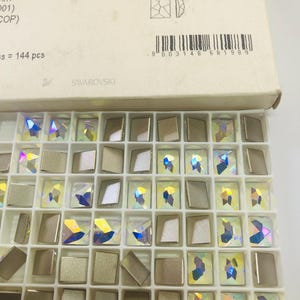 May include: A white plastic tray containing rectangular Swarovski Cosmic FB crystals, featuring a mix of iridescent, multi-colored faceted surfaces and solid matte metallic finishes. The packaging box displays the text Cosmic FB and a barcode.