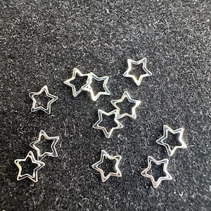 May include: A collection of silver star-shaped charms with an open center. The charms are scattered on a black surface.