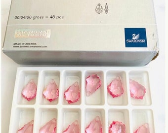 Puede incluir: Un conjunto de 12 colgantes de cristal rosa en una bandeja de plástico transparente. Los colgantes tienen forma de lágrima con un diseño en forma de estrella. La bandeja tiene 12 compartimentos, cada uno con un colgante. La caja está etiquetada con "6090 Baroque Pendant 22 x 15 mm Light Rose (223) 00/04/00 gross = 48 pcs" y "Swarovski".