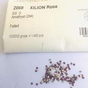 May include: A pile of small, round, amethyst-colored Swarovski crystals with silver backing. A paper label reads "SWAROVSKI ELEMENTS" and details including "XILION Rose" and "Amethyst (204)".