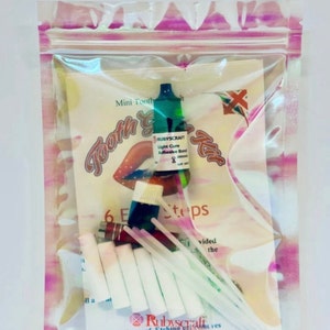 Op de afbeelding: Een doorzichtige plastic zak met een tandjuwelenset. De set bevat een klein flesje met licht uithardende lijm, een flesje met groene vloeistof, een klein rood flesje, zes witte wattenstaafjes, zes roze wattenstaafjes en twee witte tandjuwelen sjablonen. De zak heeft een roze en witte iriserende achtergrond.